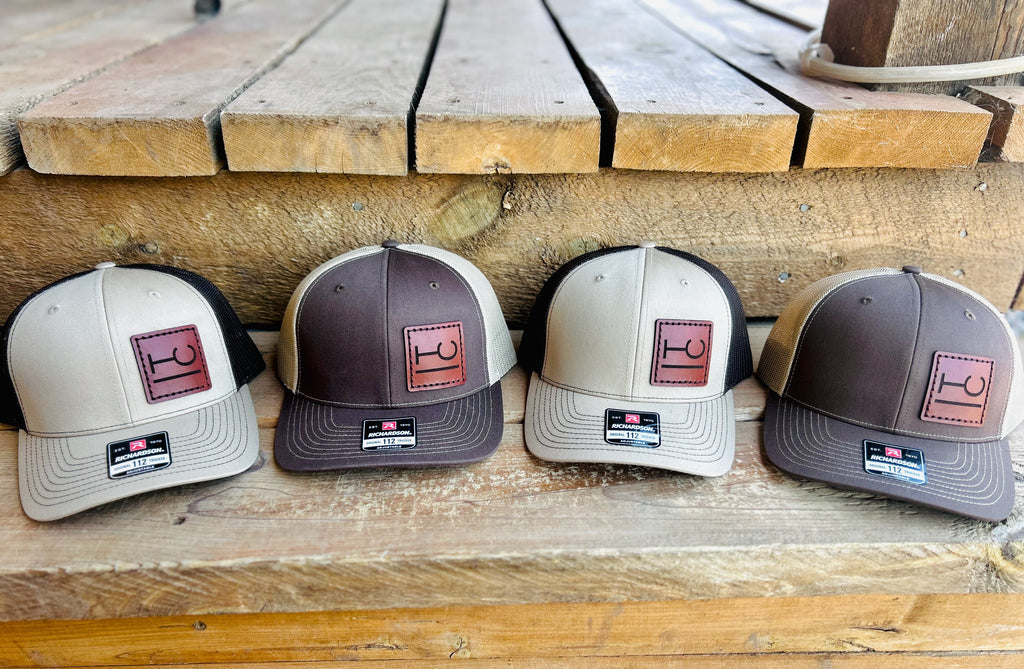 Four baseball caps with cattle brands on a leather patch displayed on a wooden surface