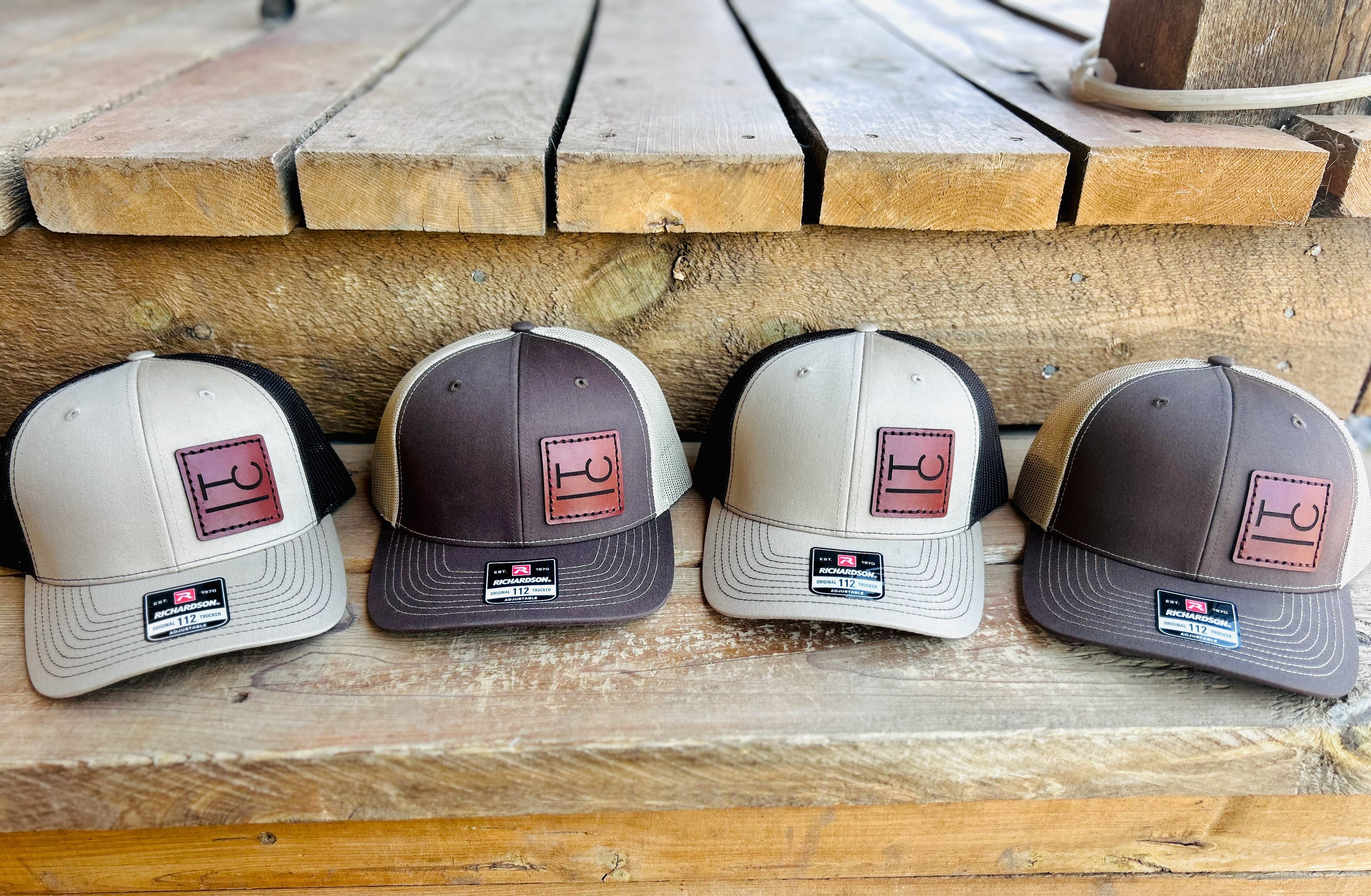 Four baseball caps with cattle brands on a leather patch displayed on a wooden surface
