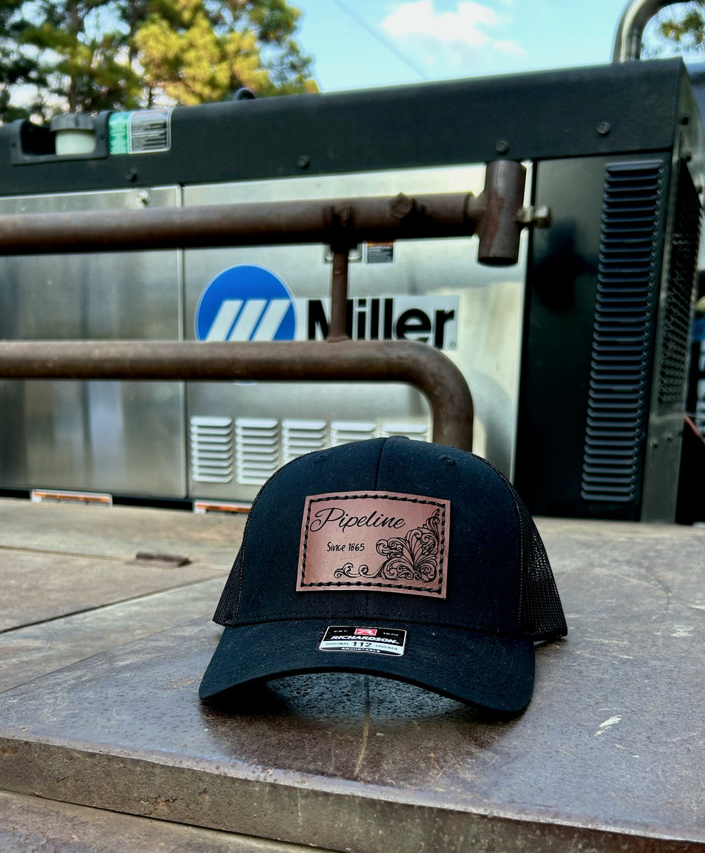 Black cap with a leather patch on a metal surface with a Miller equipment background