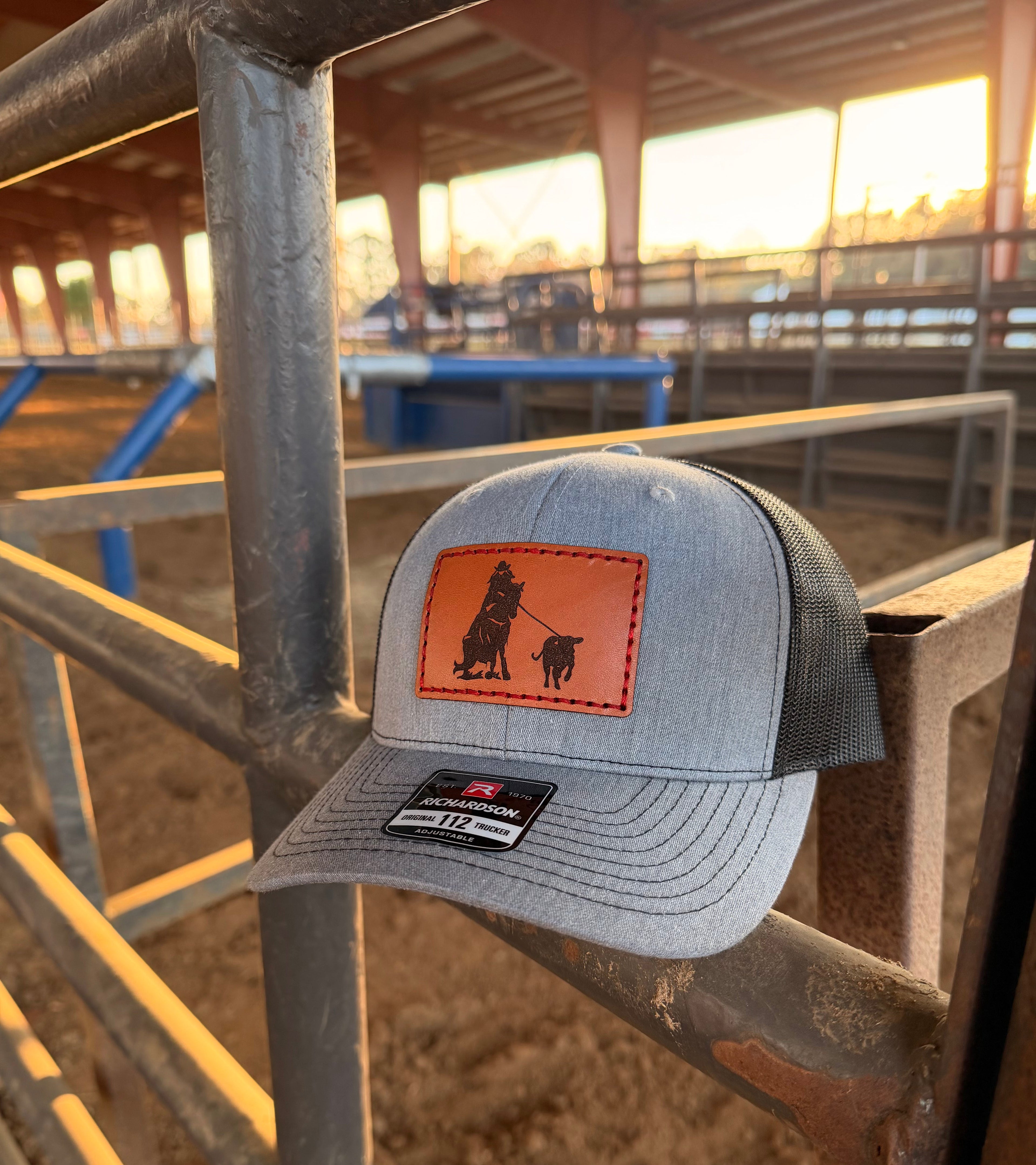 Gray cap with leather patch featuring a breakaway roping design, placed on metal bars with a barn background.