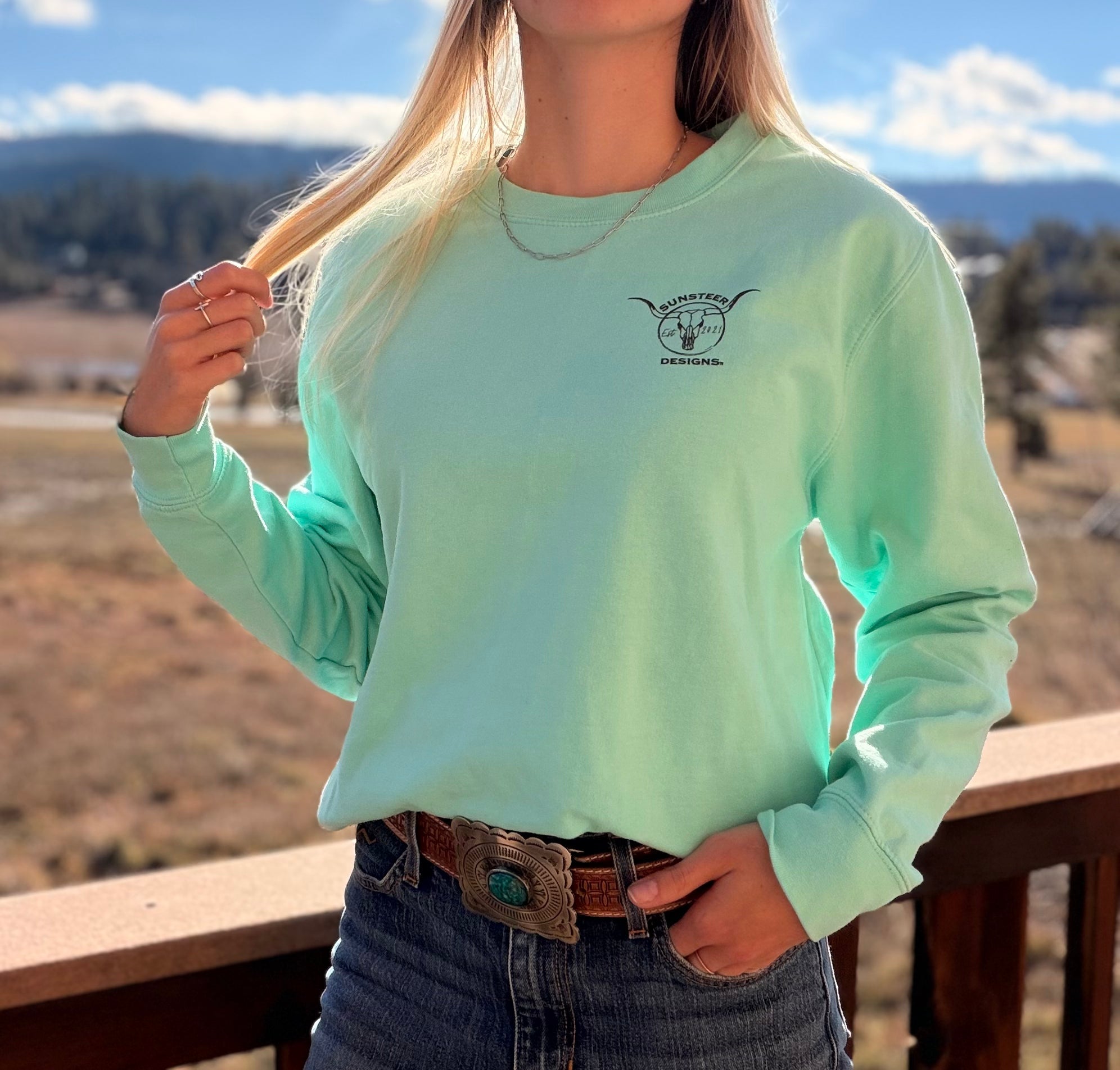Person wearing a light green long-sleeve shirt with a logo, standing outdoors with mountains in the background.