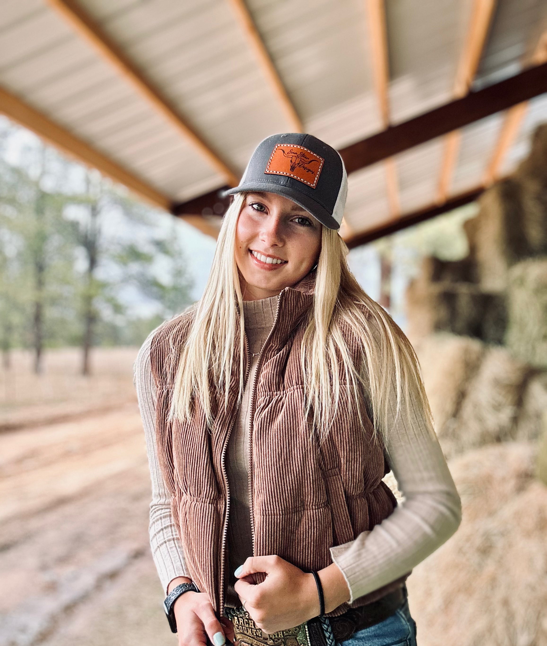 Woman wearing a brown vest and cap with a logo, standing under a wooden structure.