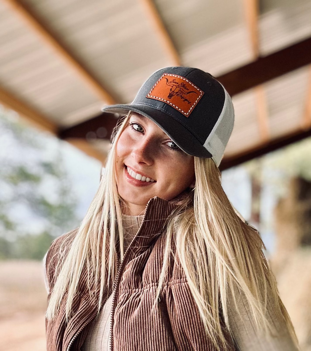 Woman wearing a brown vest and cap in an outdoor setting with trees and hay bales.