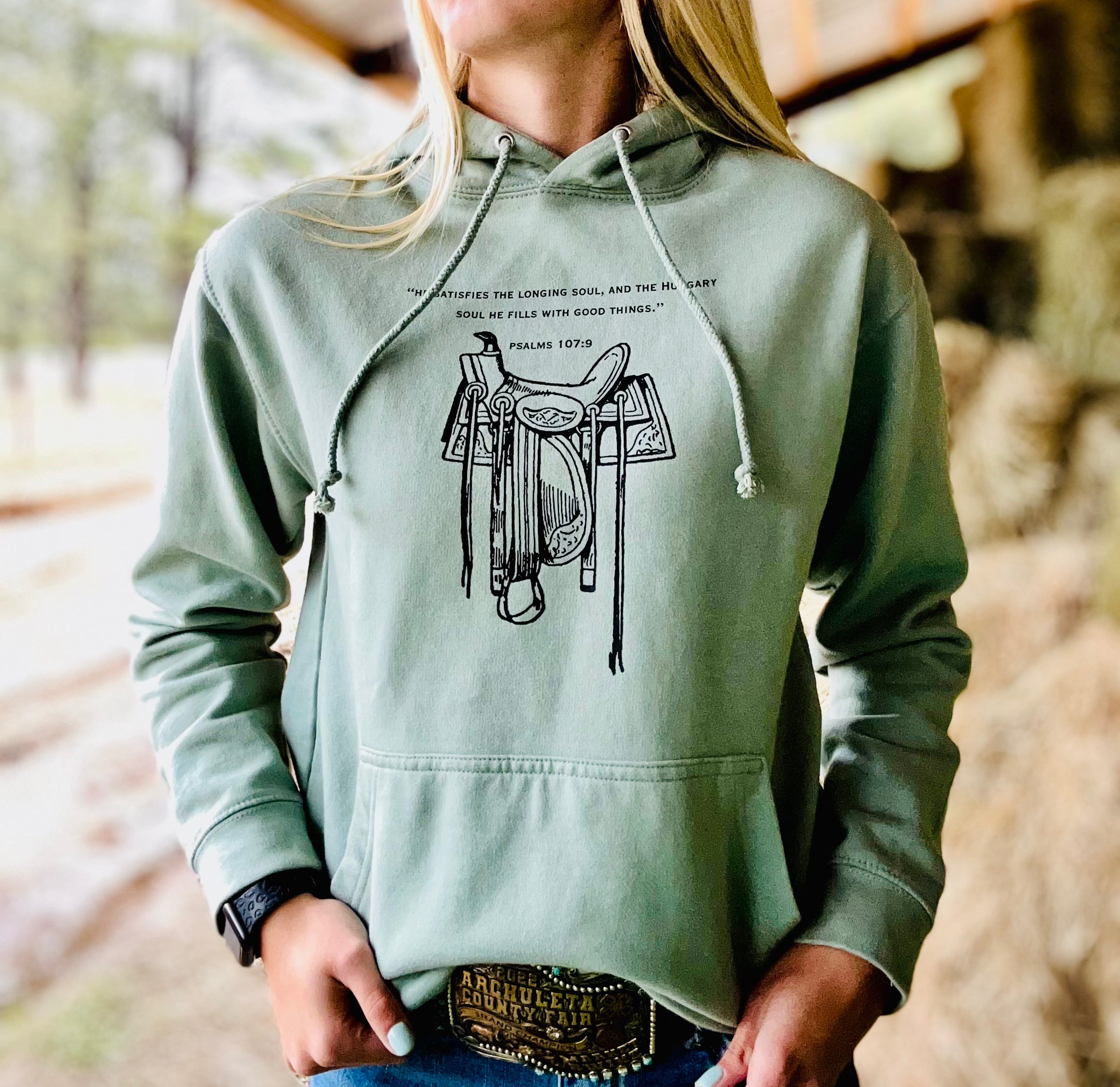 Person wearing a green hoodie with a saddle graphic and text, standing outdoors.