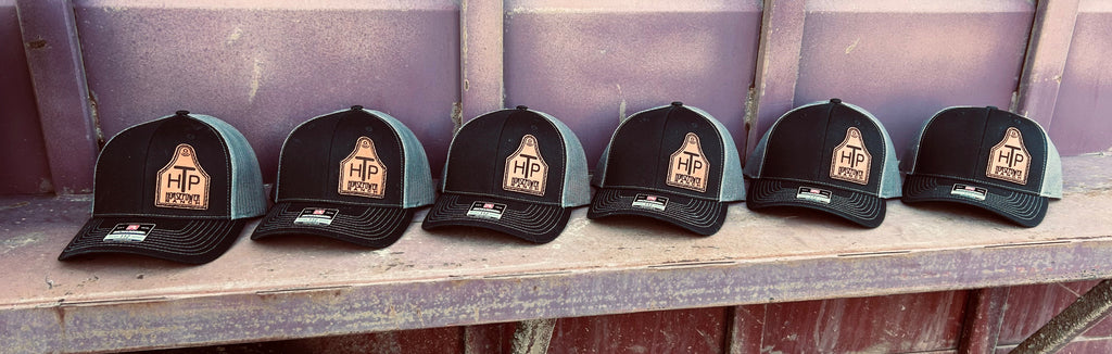 Six black and grey caps with a logo on a cowtag leather patch surface against a rustic background