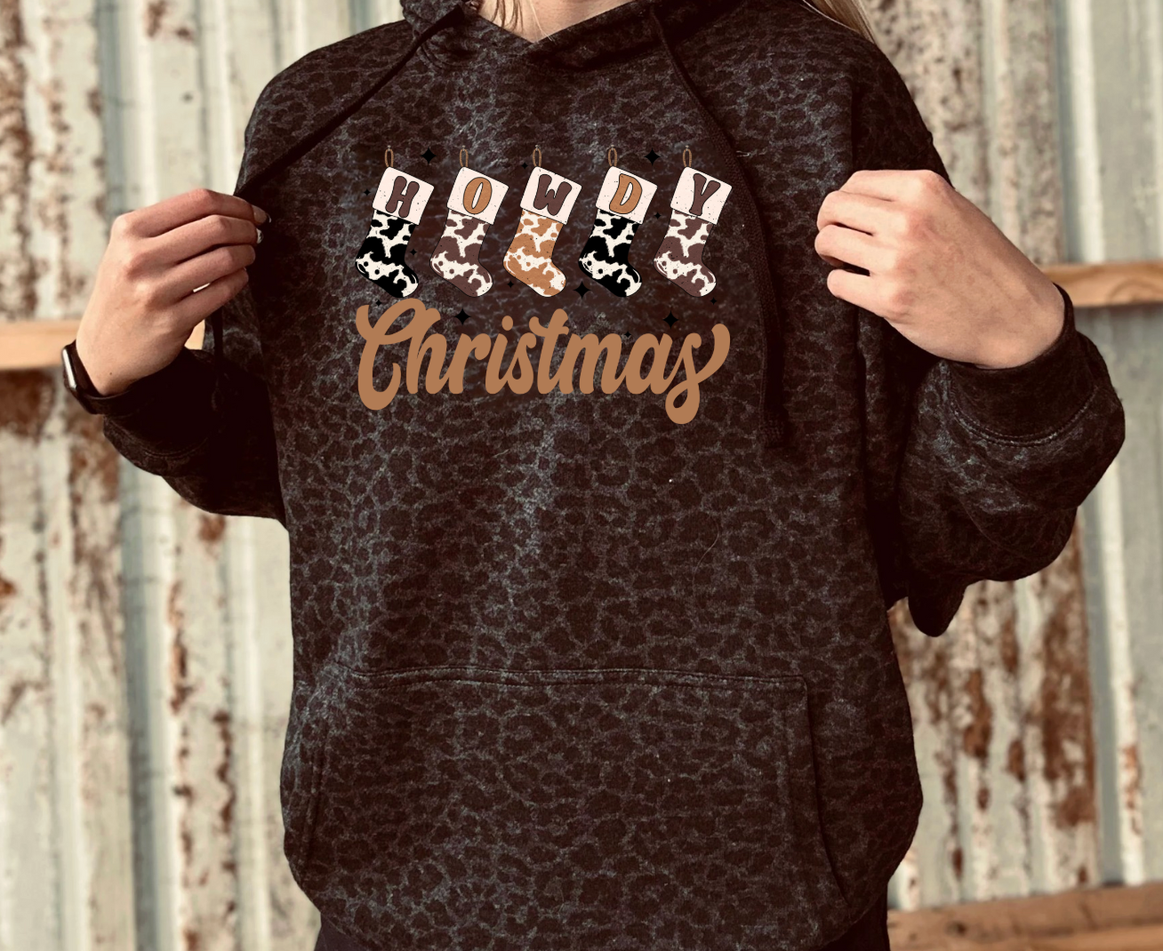 Person wearing a black leopard hoodie with 'Howdy Christmas' text and reindeer graphics against a rustic background.rd