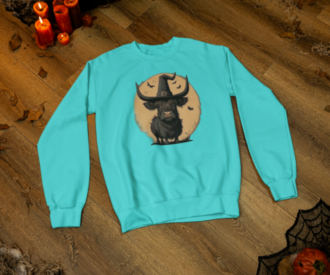 Light blue sweatshirt with a graphic design of a cow wearing a witch hat on a wooden surface.