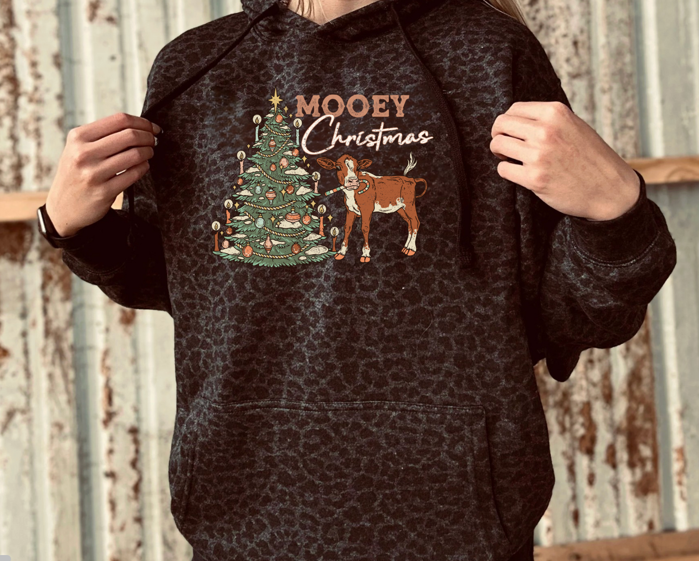 Person wearing a black leopard hoodie with a Christmas design featuring a tree and cow, against a rustic metal wall background.