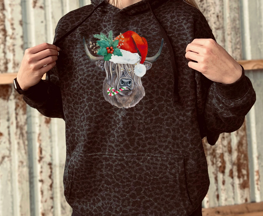 Person wearing a black leopard hoodie with a highland cow wearing a Santa hat design against a rustic metal wall background.