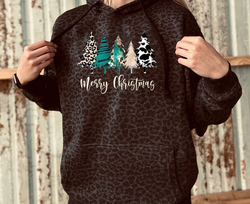 Person wearing a black leopard hoodie with Christmas tree and cow design, text 'Merry Christmas' on a rustic background.