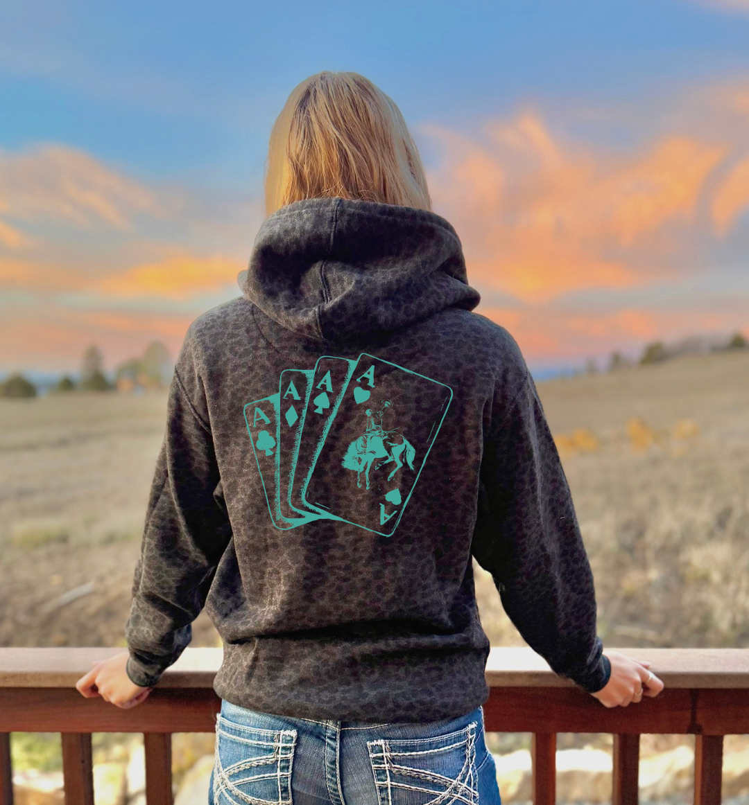 Person wearing a black leopard hoodie with a aces bronc cards design on the back, standing on a wooden deck with a scenic view of mountains and clouds.