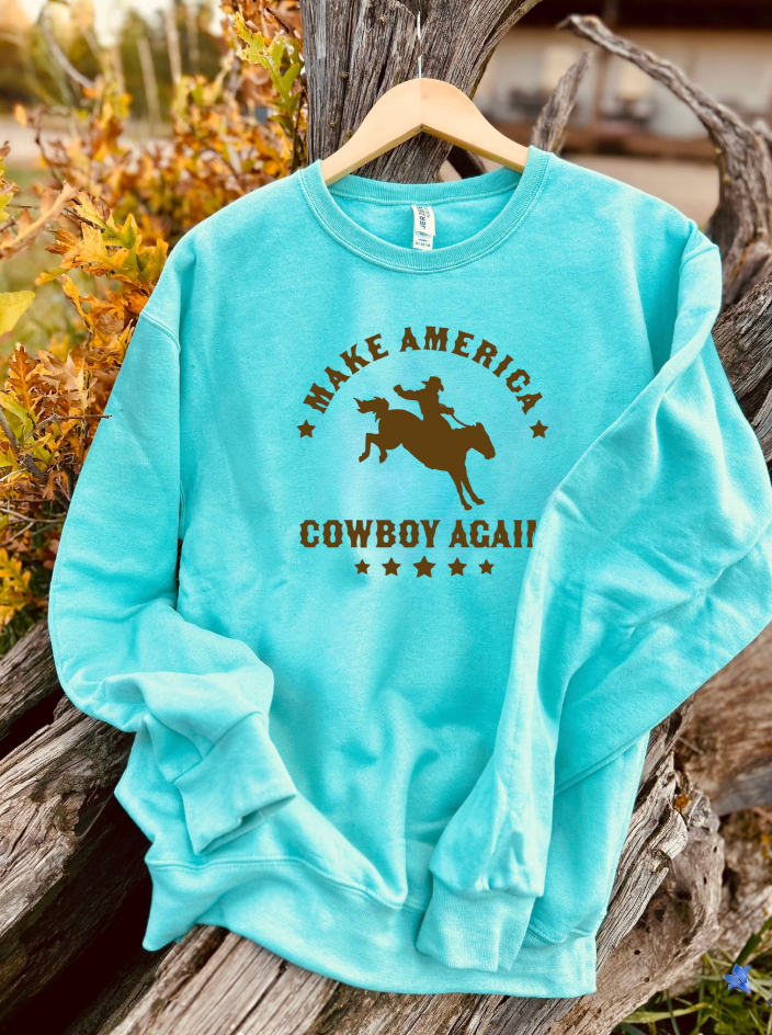 Mint sweatshirt with brown 'Make America Cowboy Again' text and graphic on a wooden surface.