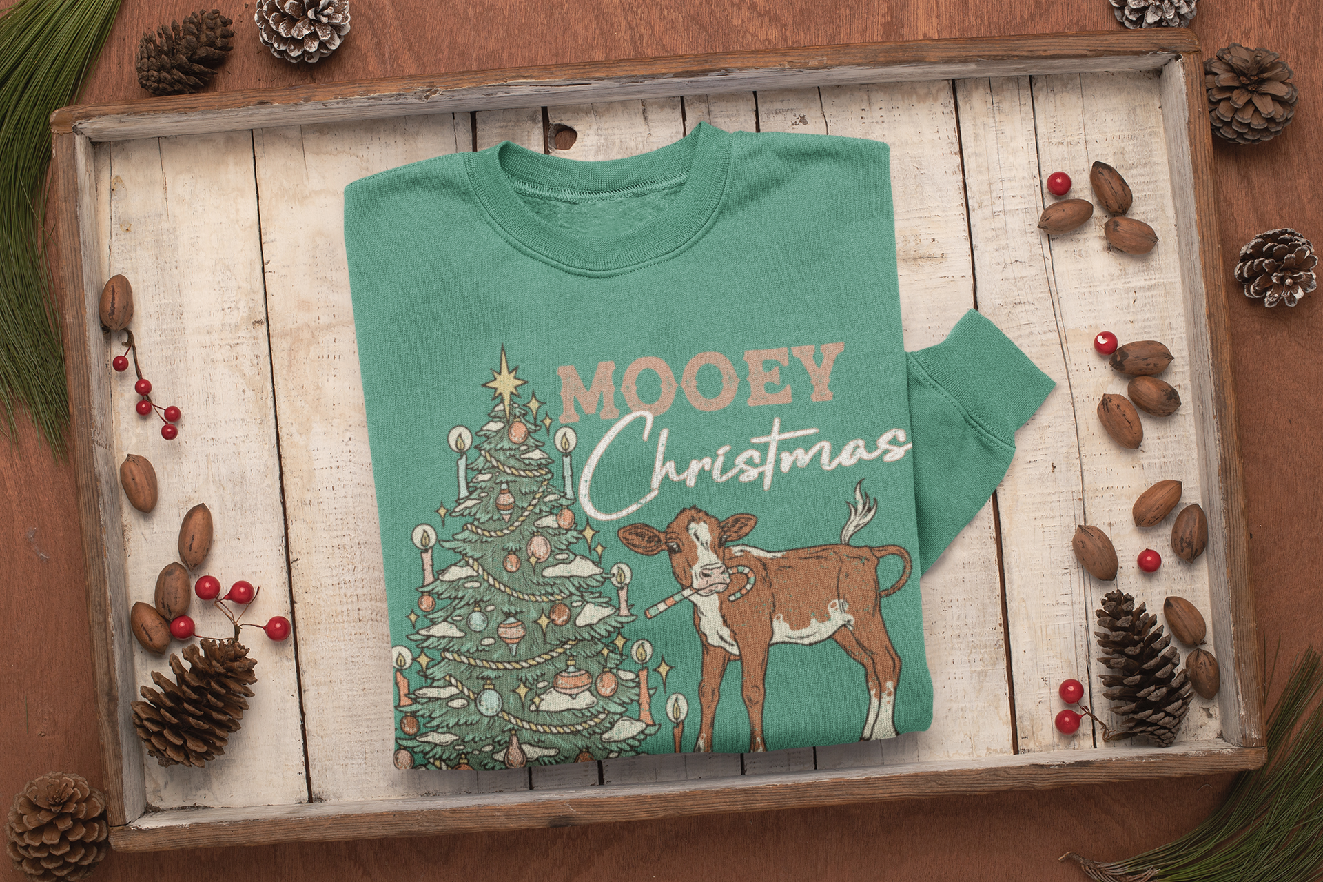 Green sweatshirt with Christmas design featuring a tree and a calf on a wooden surface with pinecones and berries.