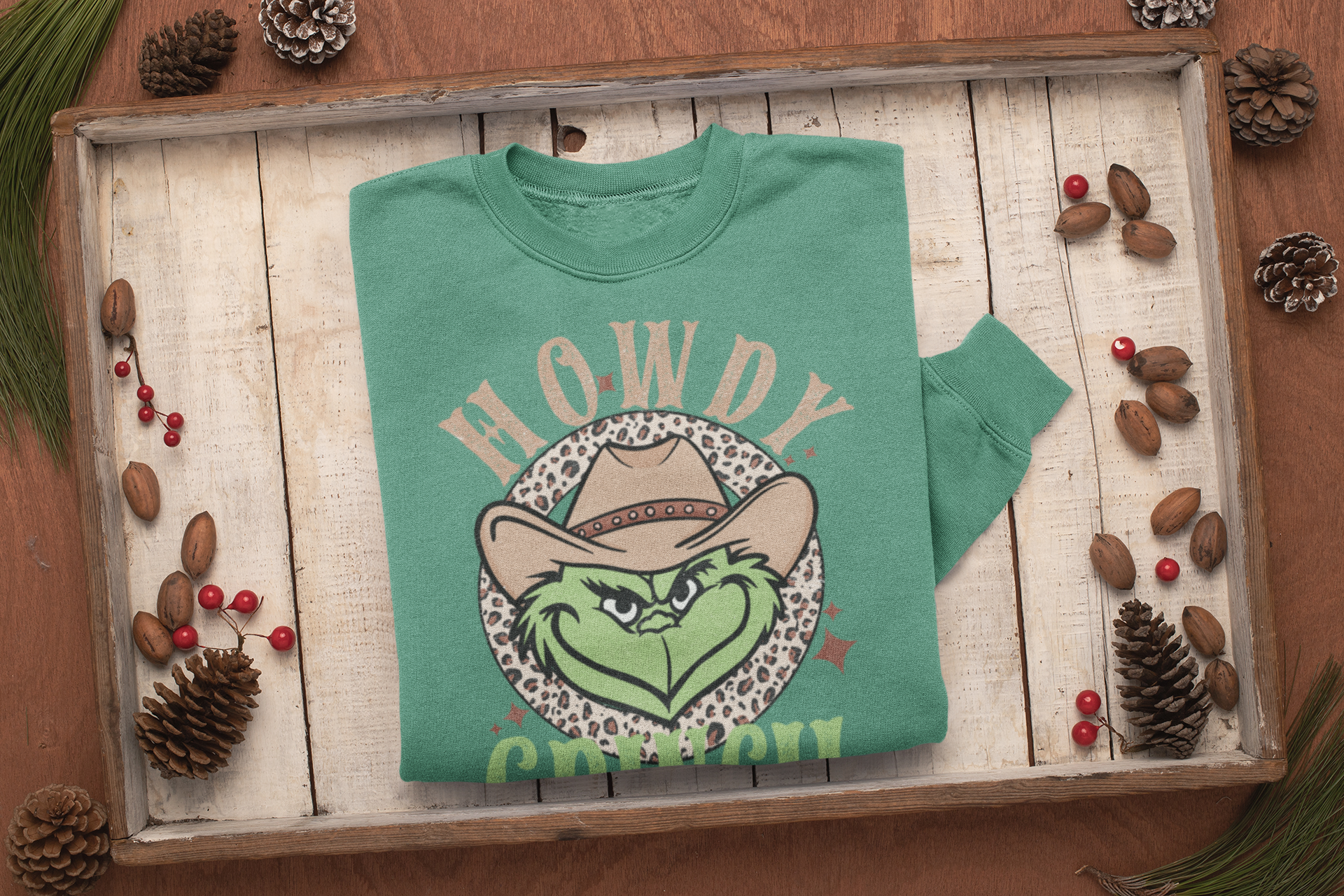 Green sweatshirt with 'Howdy Cowboy' design on a wooden surface with pinecones and berries.